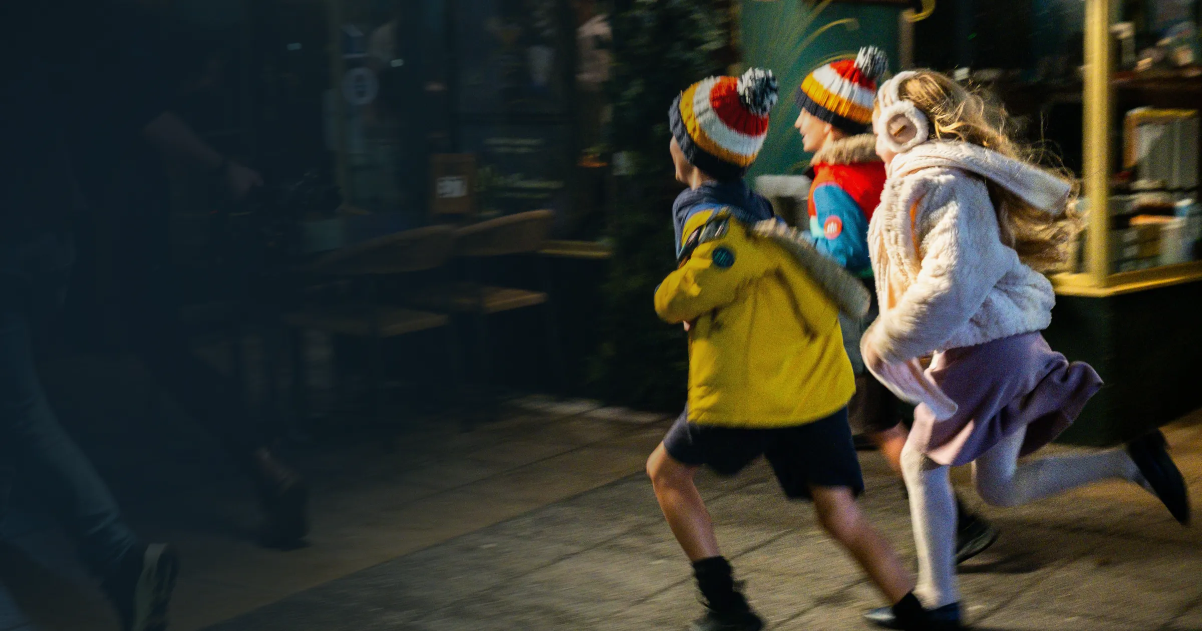 Three children in winter clothing, wearing colourful hats, run energetically on a city street at night. A camera crew follows closely, capturing their joyful motion.